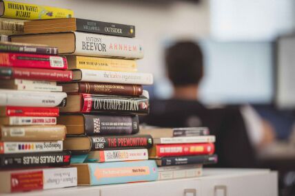 Three uneven stacks of books  of multiple sizes and colours rests on a table, with a person seated in front out of focus looking at a computer monitor.